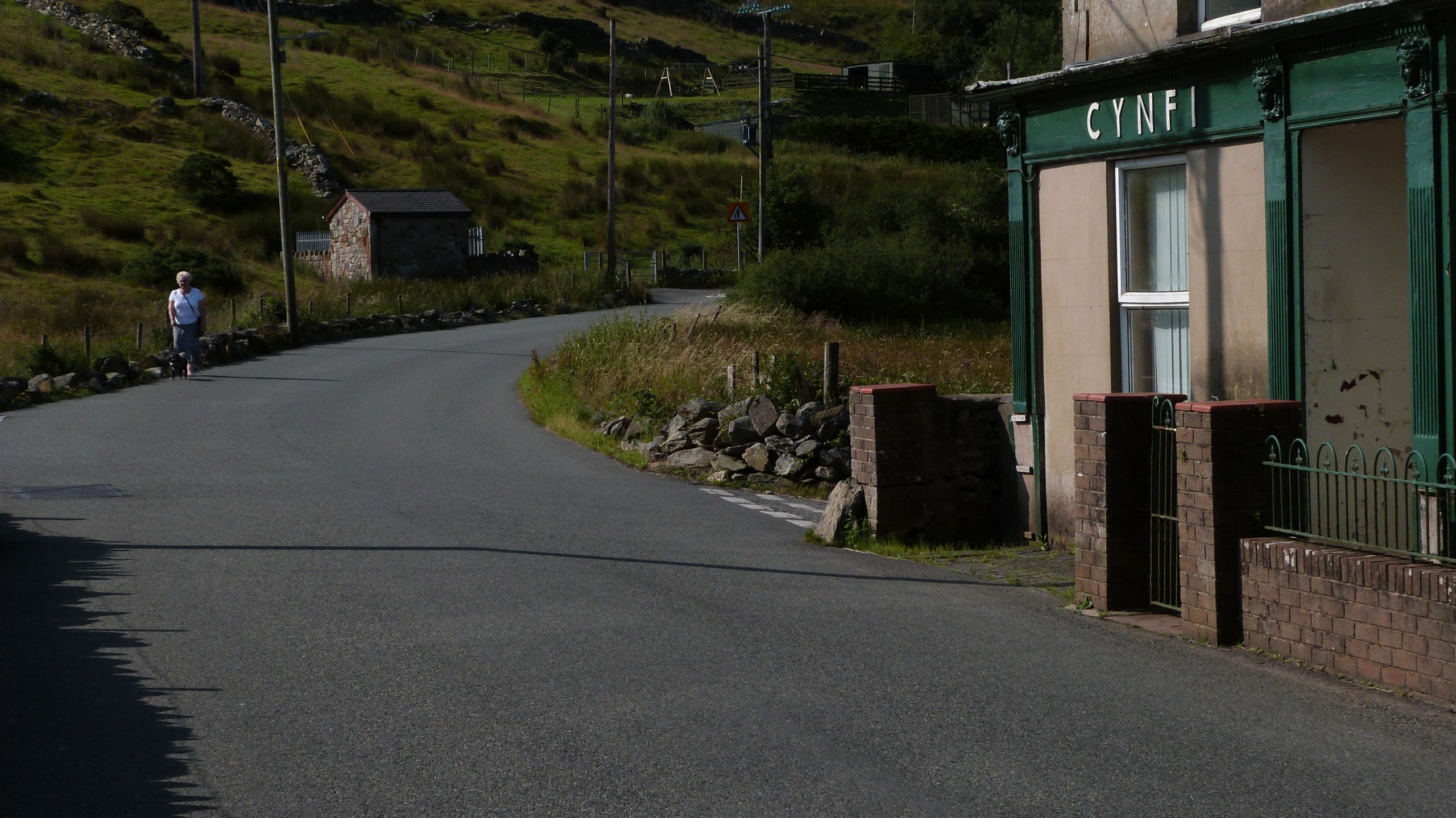 Pensiynwraig yn cerdded i lawr stryd wag, tuag at hen siop wedi cau 