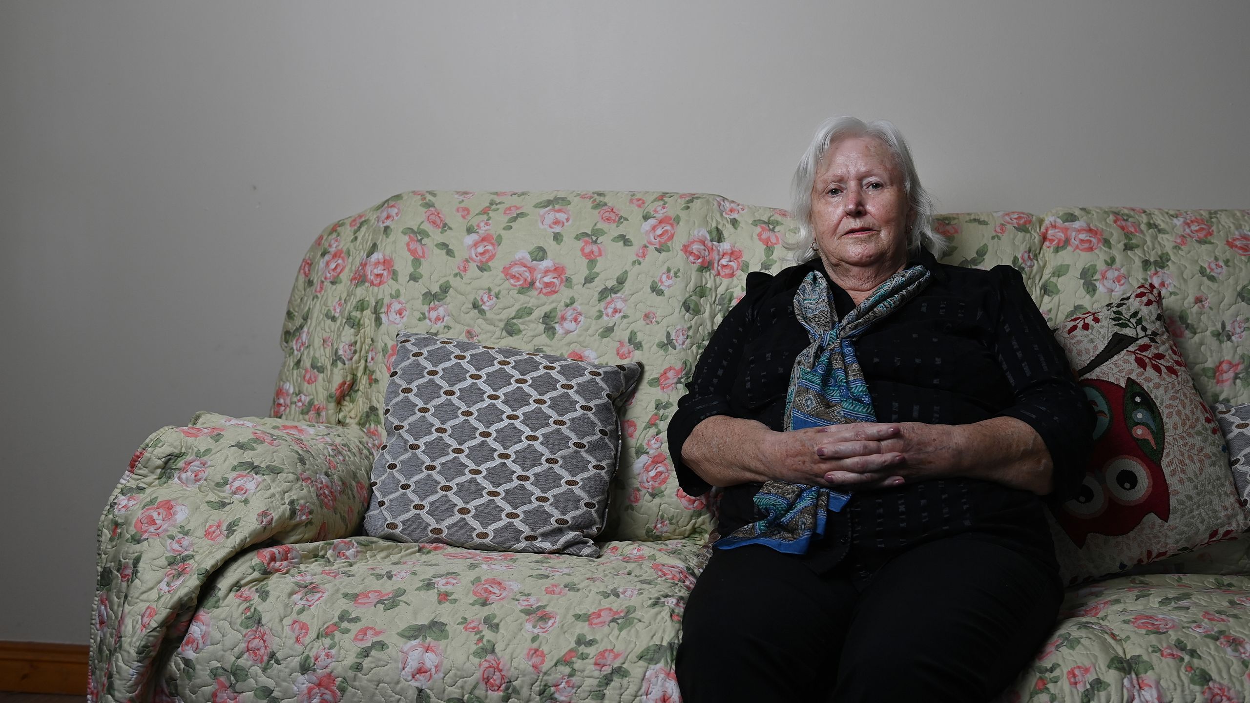 Mary Moriarty, wearing a dark blue outfit and patterned neck scarf, sitting on a cream coloured sofa. She is an older woman with shoulder length white hair.