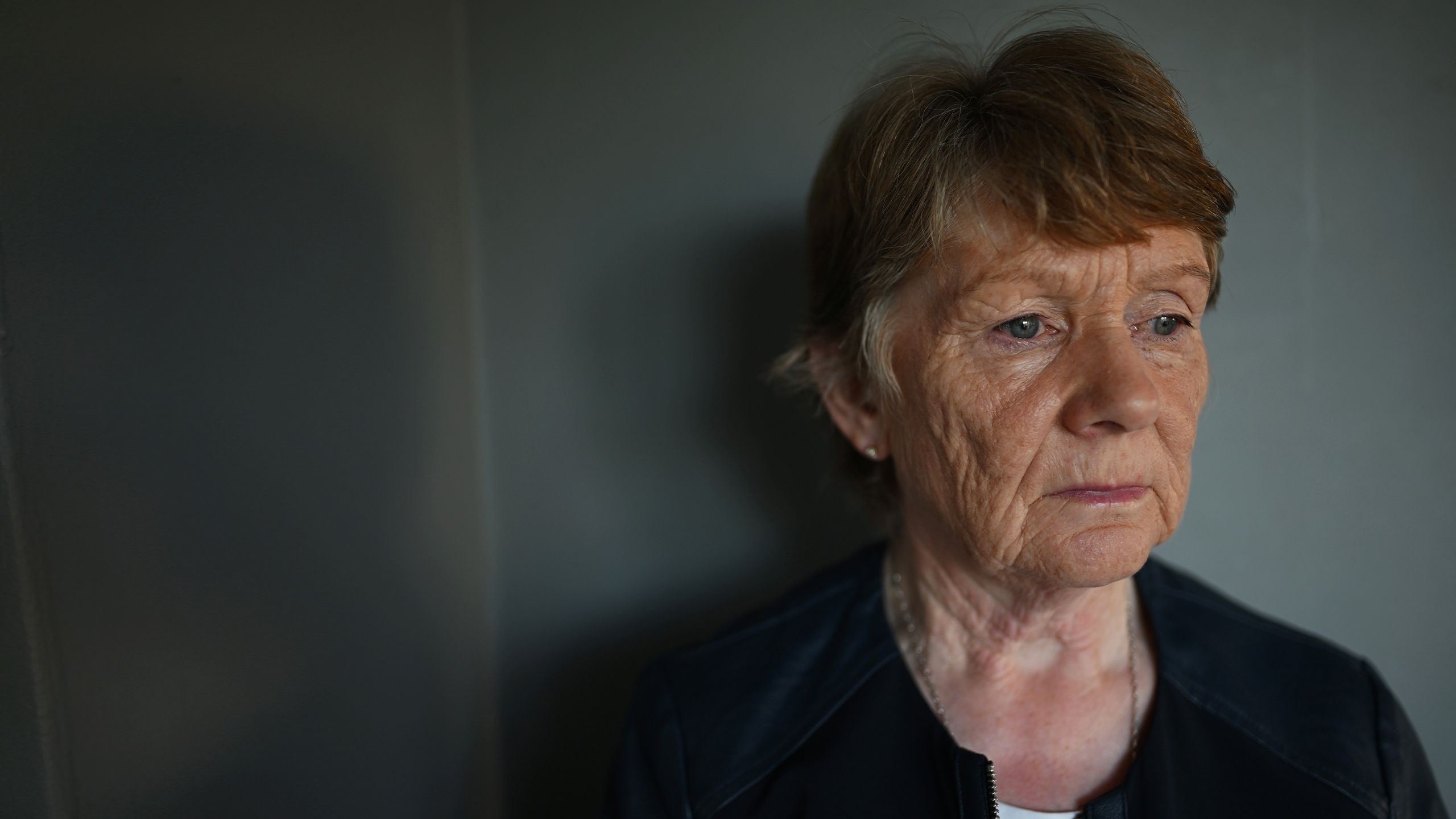 Catherine Corliss pictured against a grey wall. She is an older woman with short cropped brown hair and she is looking to the right off camera, wearing a black top.