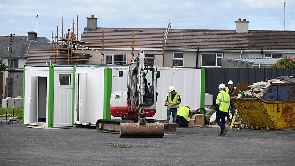 Image showing workers at the site.  There are several people pictured in hi-visability clothing, with a digger in the foreground and a skip to the right.  White and green mobile cabins are are pictured in the background.