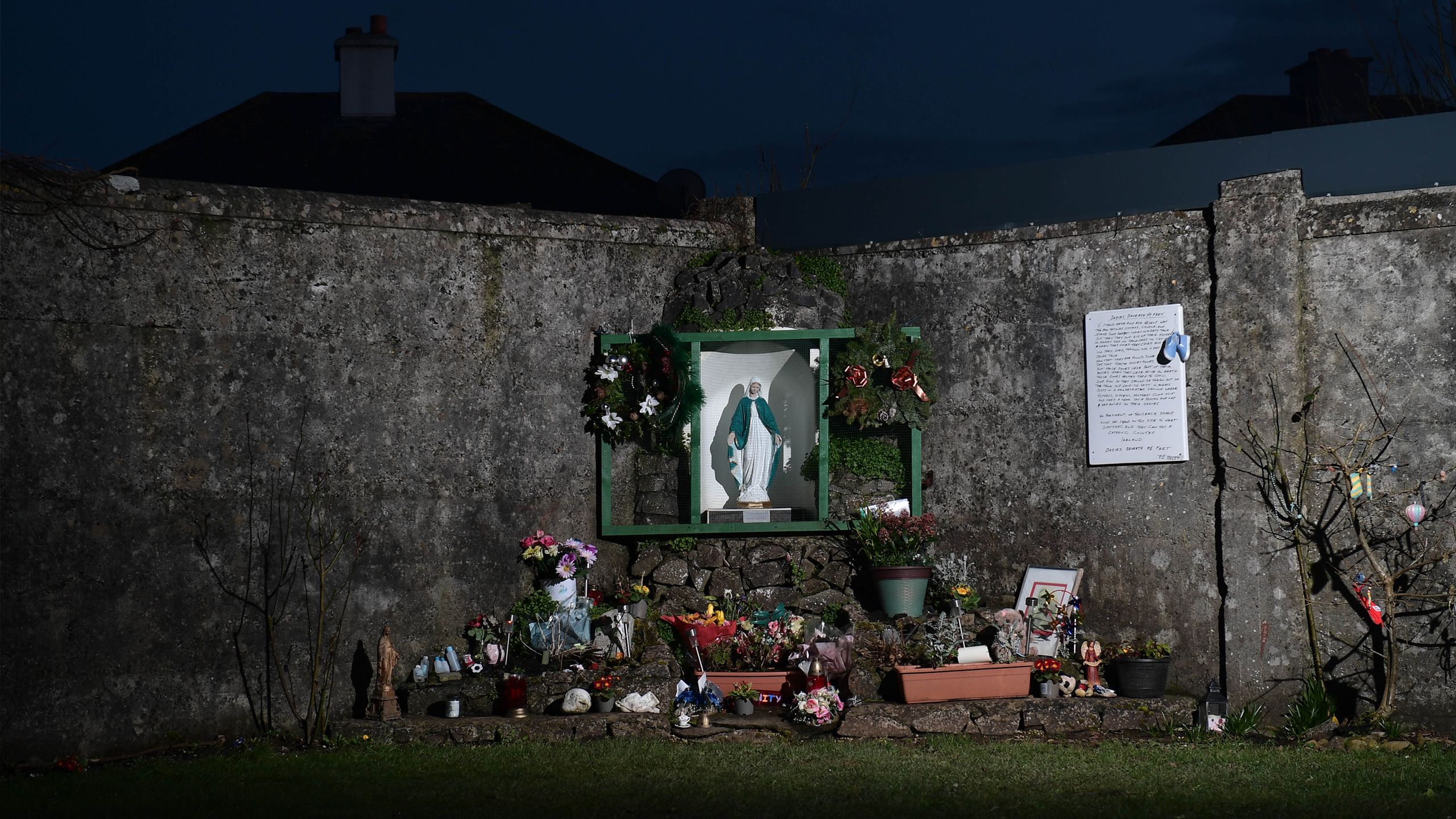 A night time picture of the memorial at Tuam. It shows a statue in the background surrounded by smaller memorials on the ground.