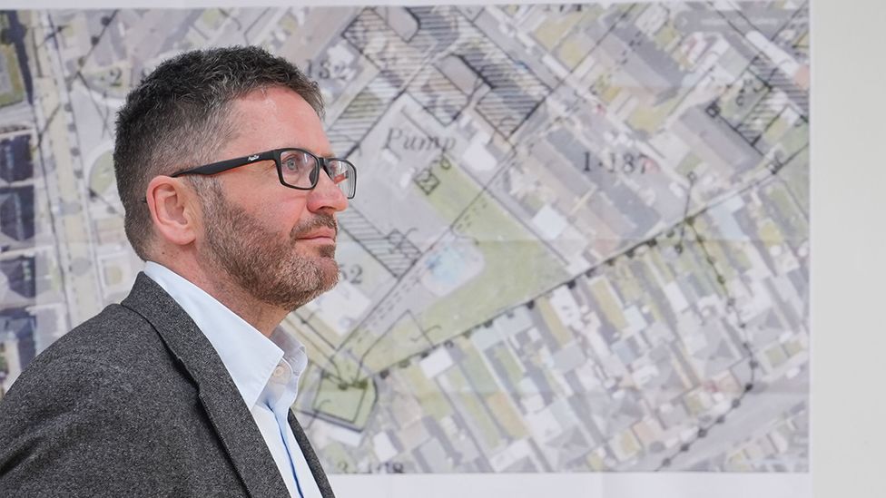 Daniel MacSweeney, a man with glasses, wearing a grey suit jacket and light shirt, is pictured in front of a large map showing construction blueprints