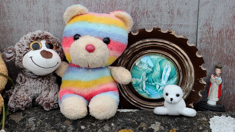 Soft toys and a framed picture left on the site as a memorial.  There is a brown teddy bear on the left with big eyes, beside it a larger rainbow reddy bear, a framed picture of a statue followed by a soft toy seal sitting on the ground.