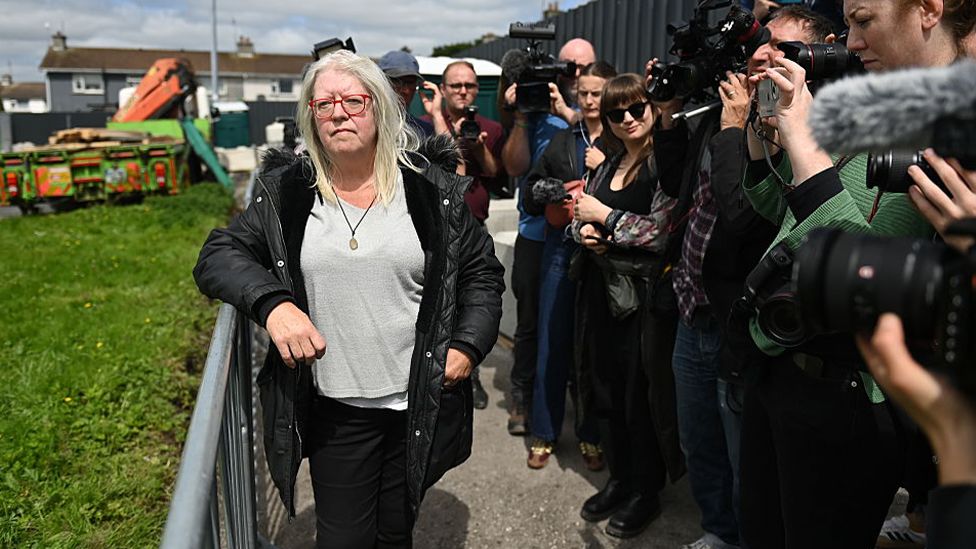 Anna Corrigan standing against a temporary barrier at the site in Tuam.  She is surrounded by photographers and media crews.