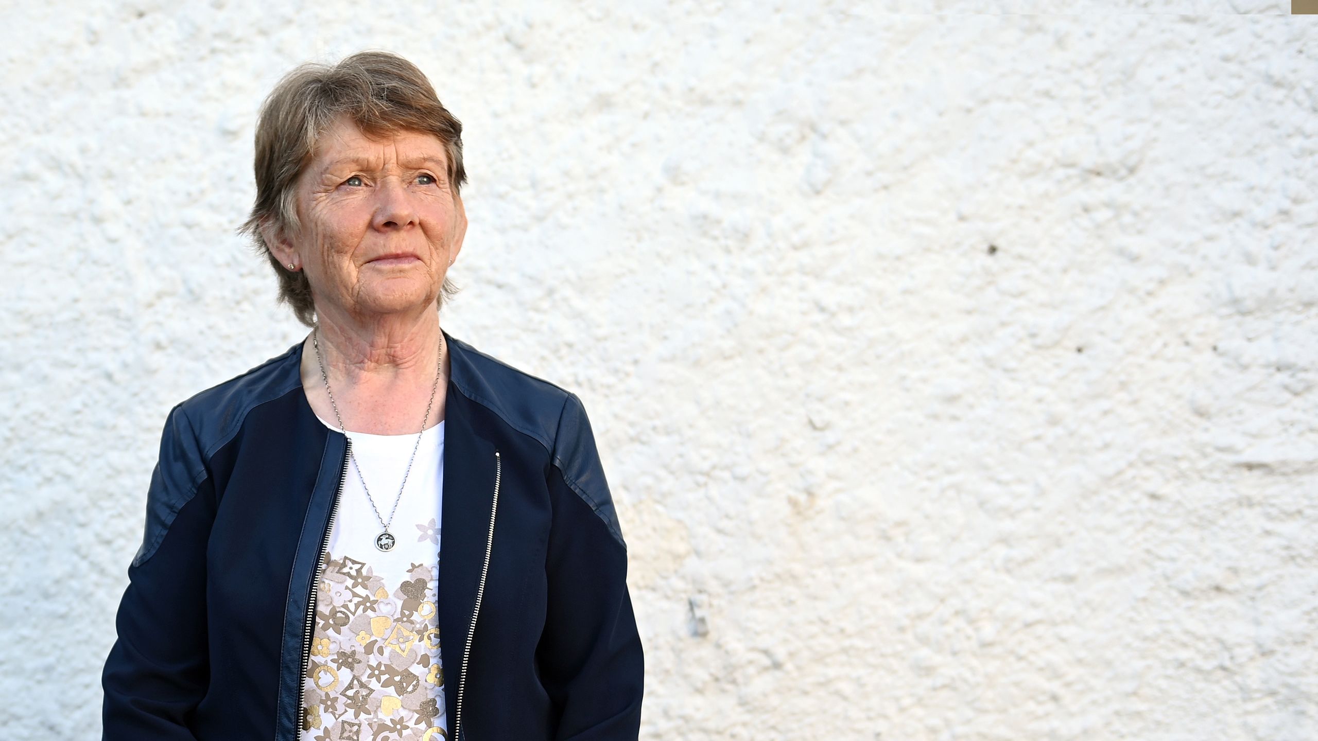Catherine Corliss, wearing a blue jacket, standing looking to the right. She is pictured against a white wall