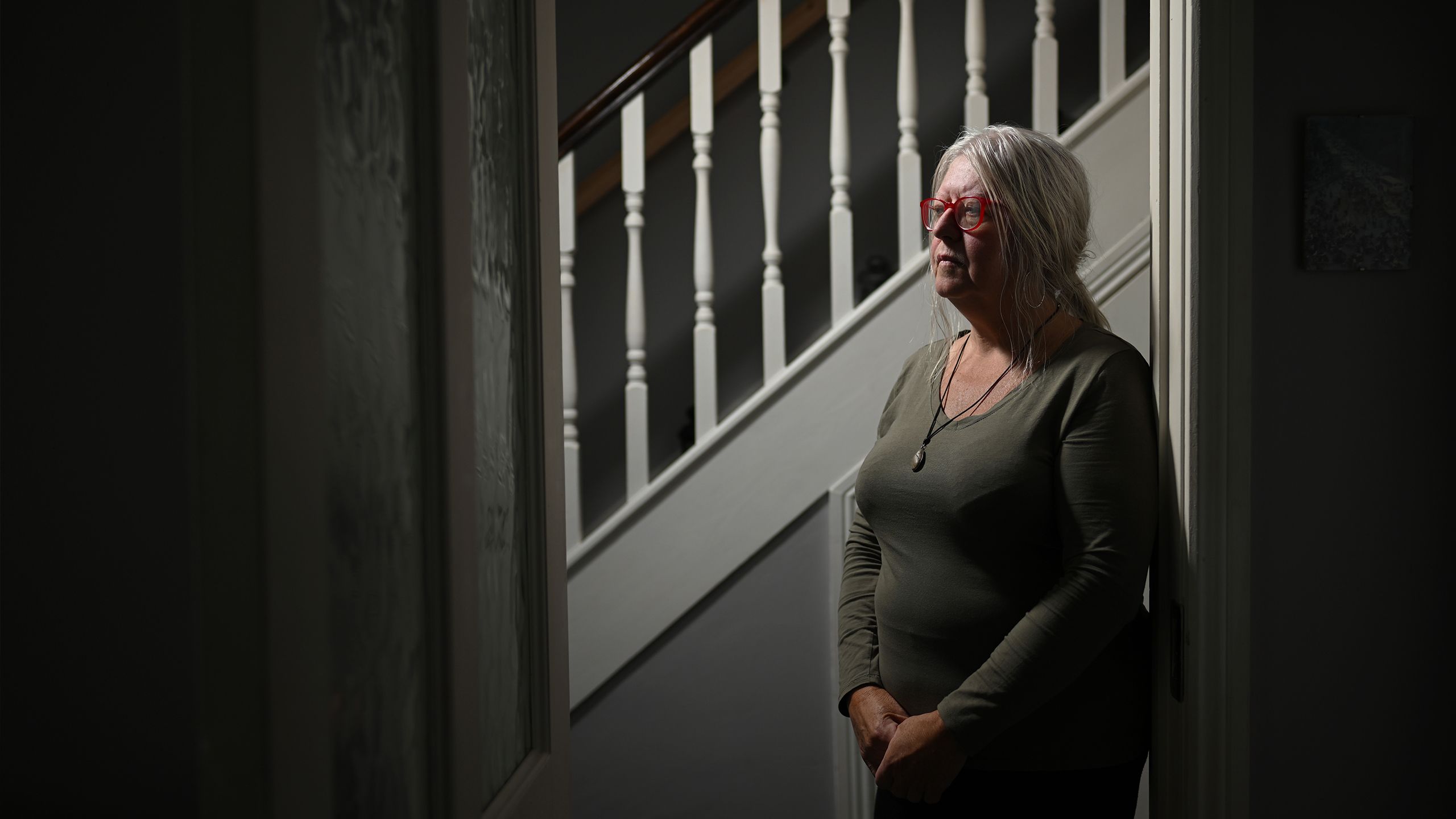 Anna Corrigan pictured in low light in front of a staircase. She is an older woman with long white hair, wearing a green top looking off to the left of camera.