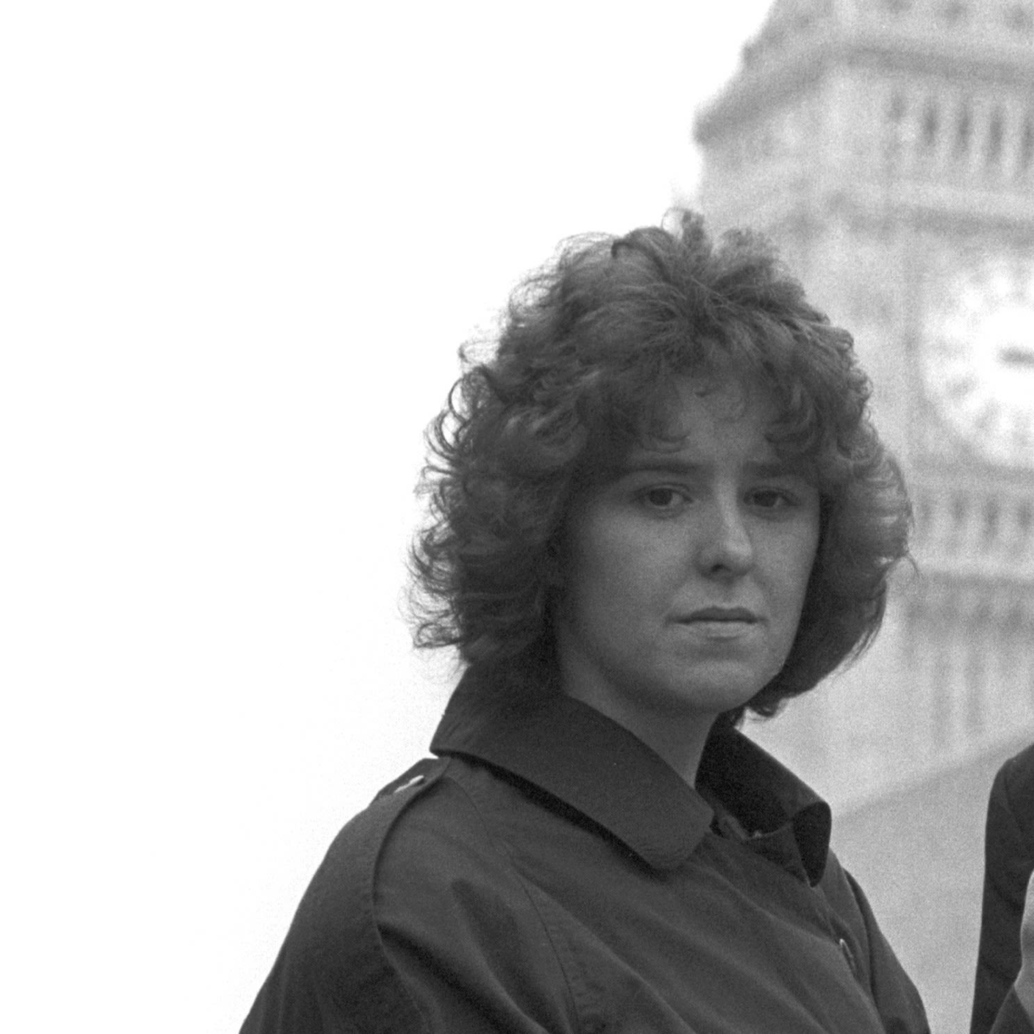 A black and white image of a woman in front of Big Ben looking down the lens. She has brown permed hair and is looking stern