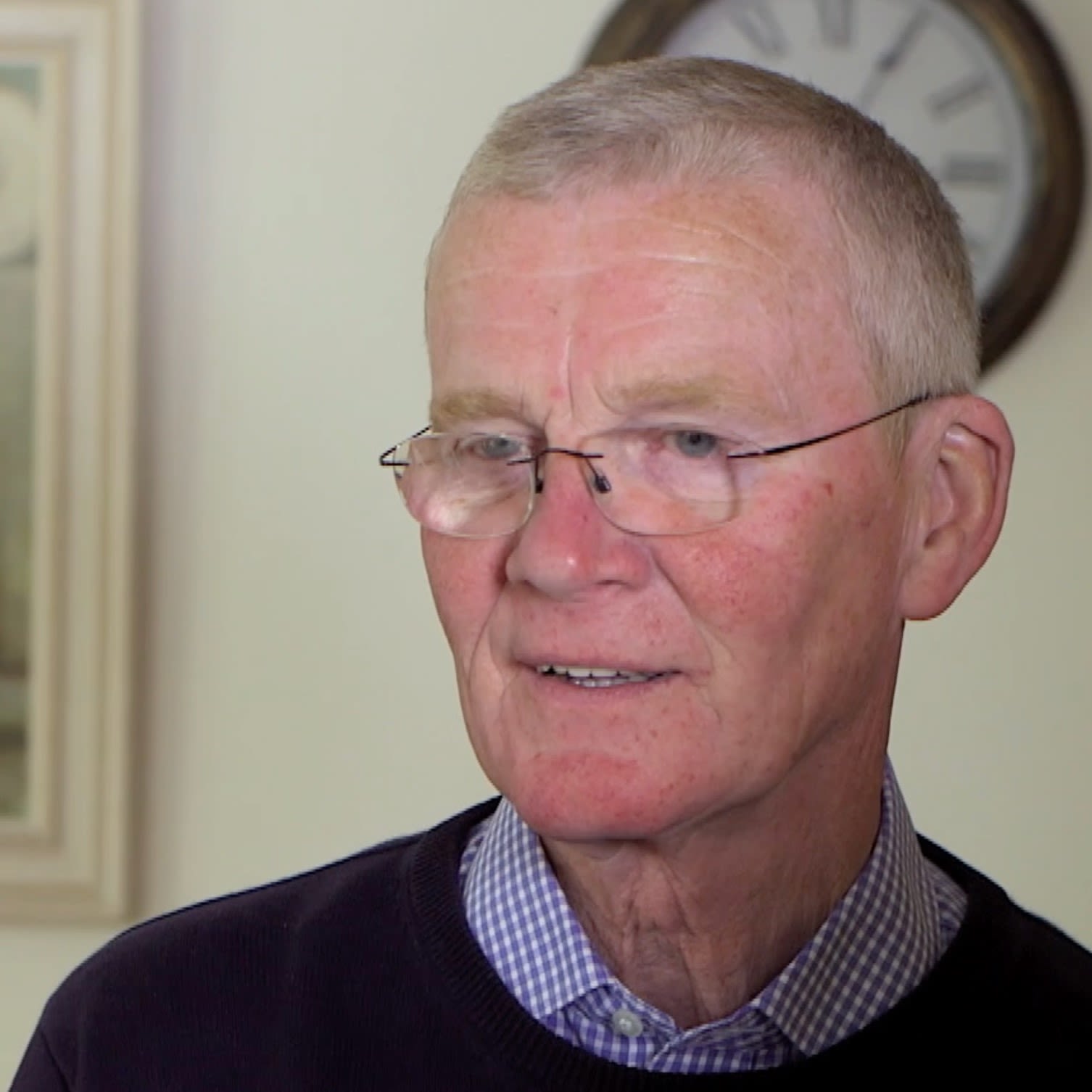 Frank Foley is looking past the camera. He is wearing glasses and his shoulders are visible, wearing a blue chequered shirt under a black jumper. There is a clock on the wall behind him.