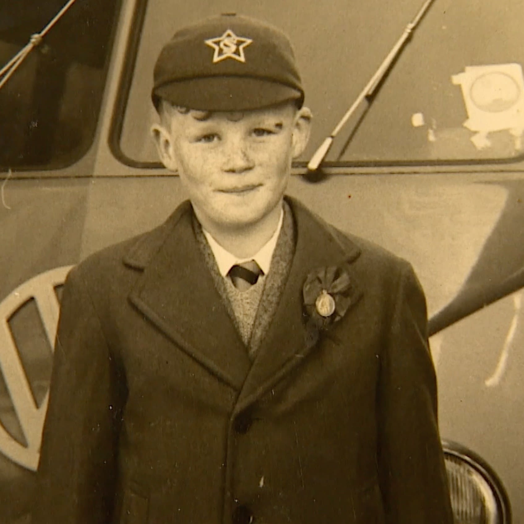 A sepia image of Frank as a young boy. He is wearing a school cap and blazer and is stood in front of a VW campervan.