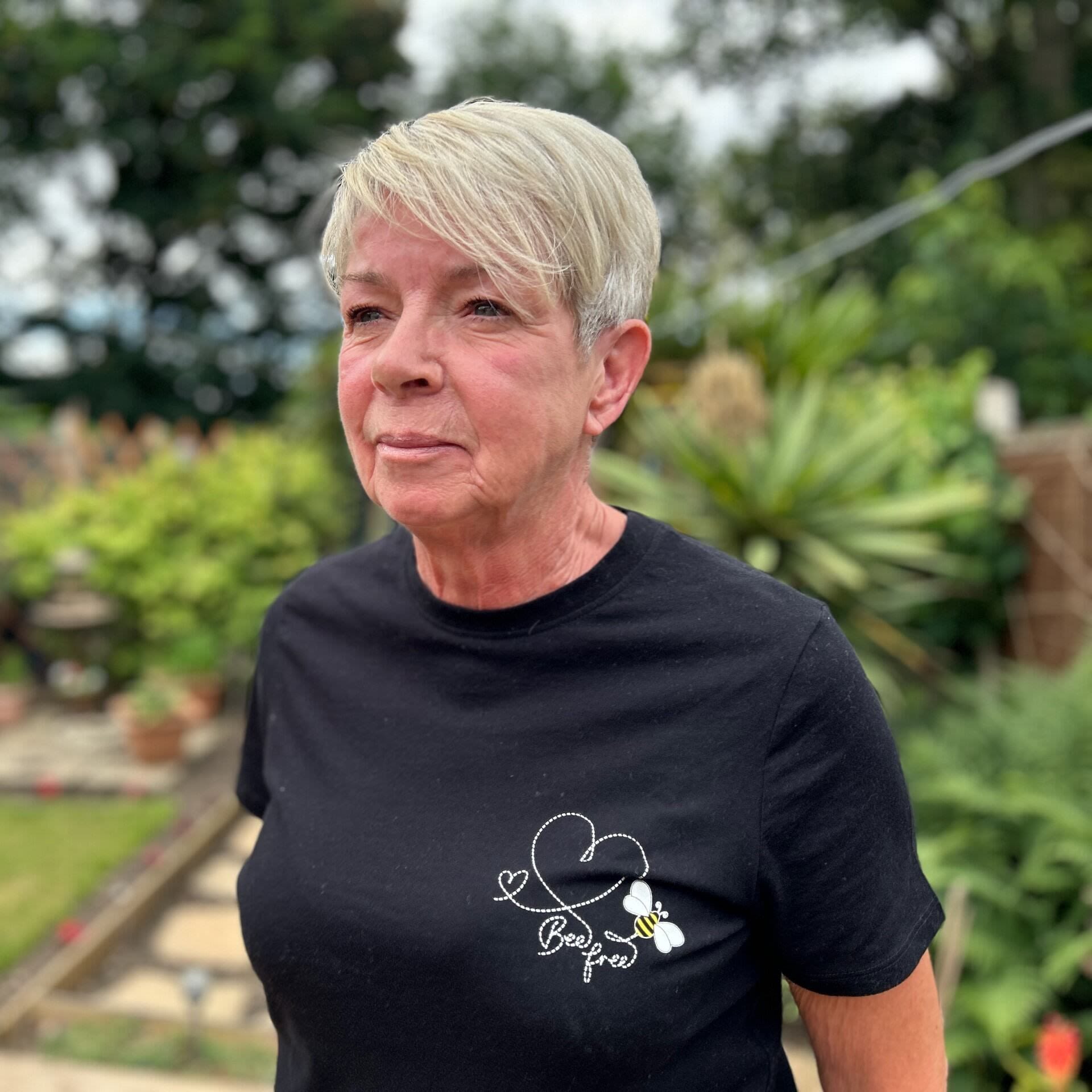Maureen Mitchell, a woman in a black top and short blond hair, is stood looking past the camera. In the backgroud is a blurred background.