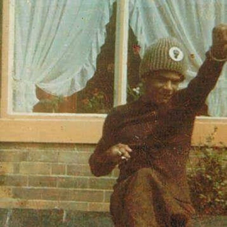 A grainy picture from the early 1970s of a young man Paul Anthony Davies performing a karate kick. he's wearing a green knitted hat with the black power logo on it and is standing in front of a house with net curtains in the window