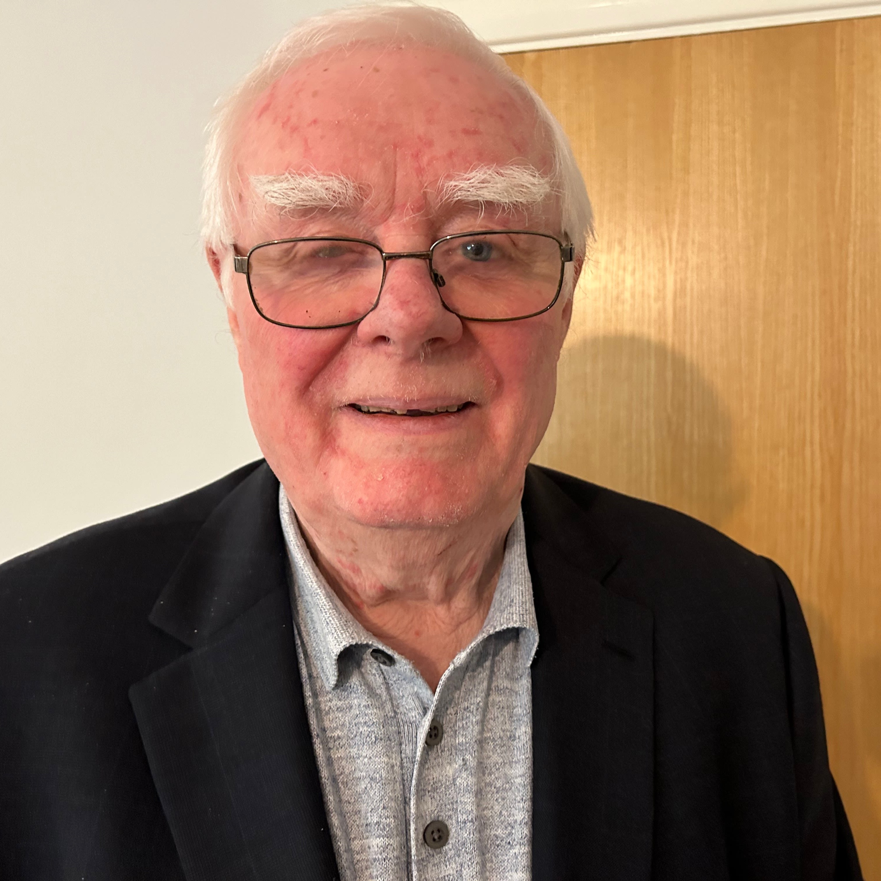 Billy Power is looking into the camera, smiling slightly. He is wearing a grey shirt and black jacket, with glasses, and has white hair.