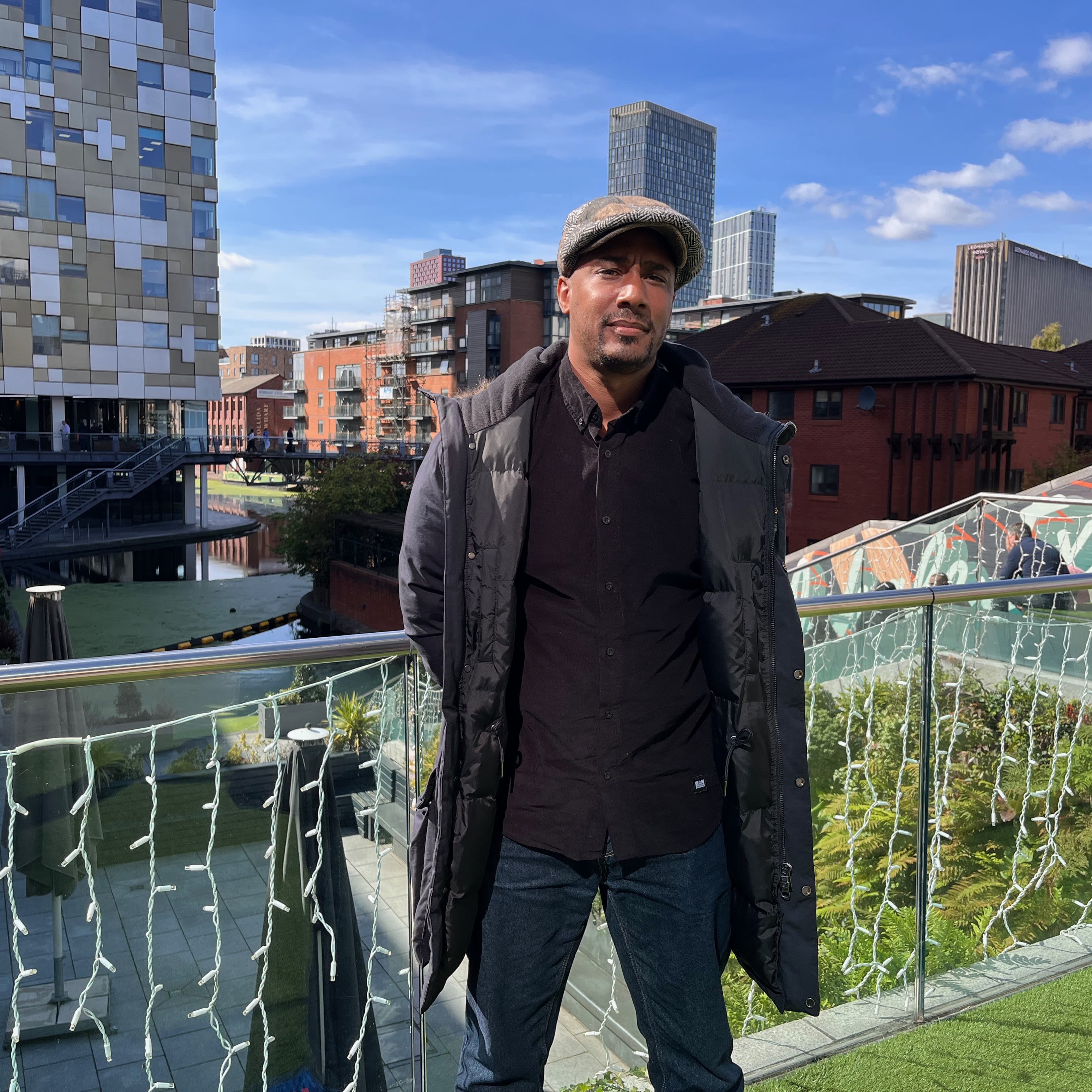 Paul Bridgewater is standing on a balcony. He is wearing a flat cap, a knee-length black coat, jeams and a dark jumper. Behind him is a blue sky, various buildings and a glimpse of the Birmingham canals.
