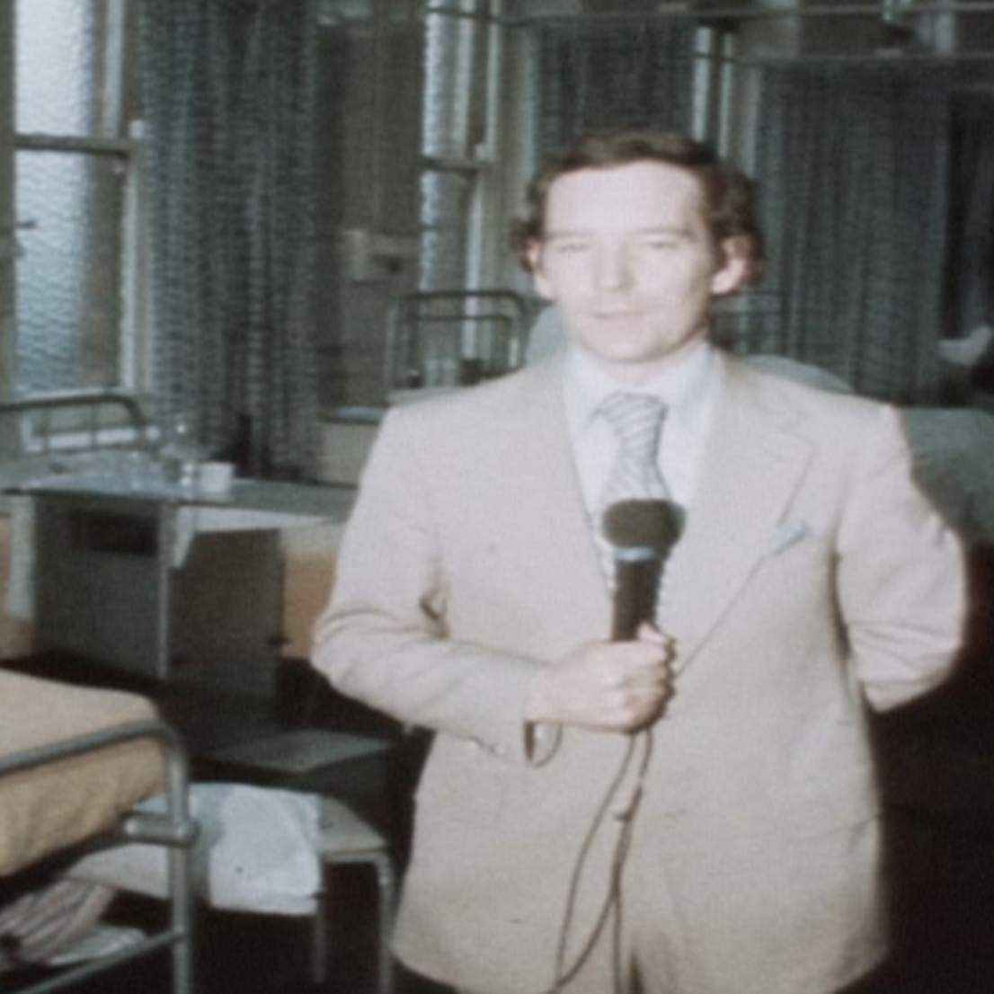 Old picture of Michael Buerk in a hospital. He is wearing a pale suit and holding a microphone. Behind him are hospital beds with yellow sheets and windows.