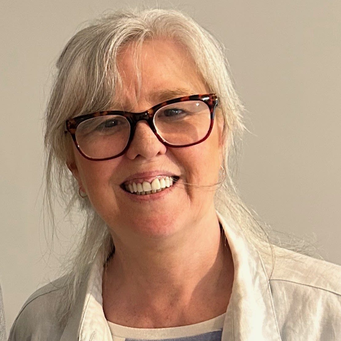 a colour picture of a woman in glasses, late 50s, with silvery hair. She's smiling against a plain white wall