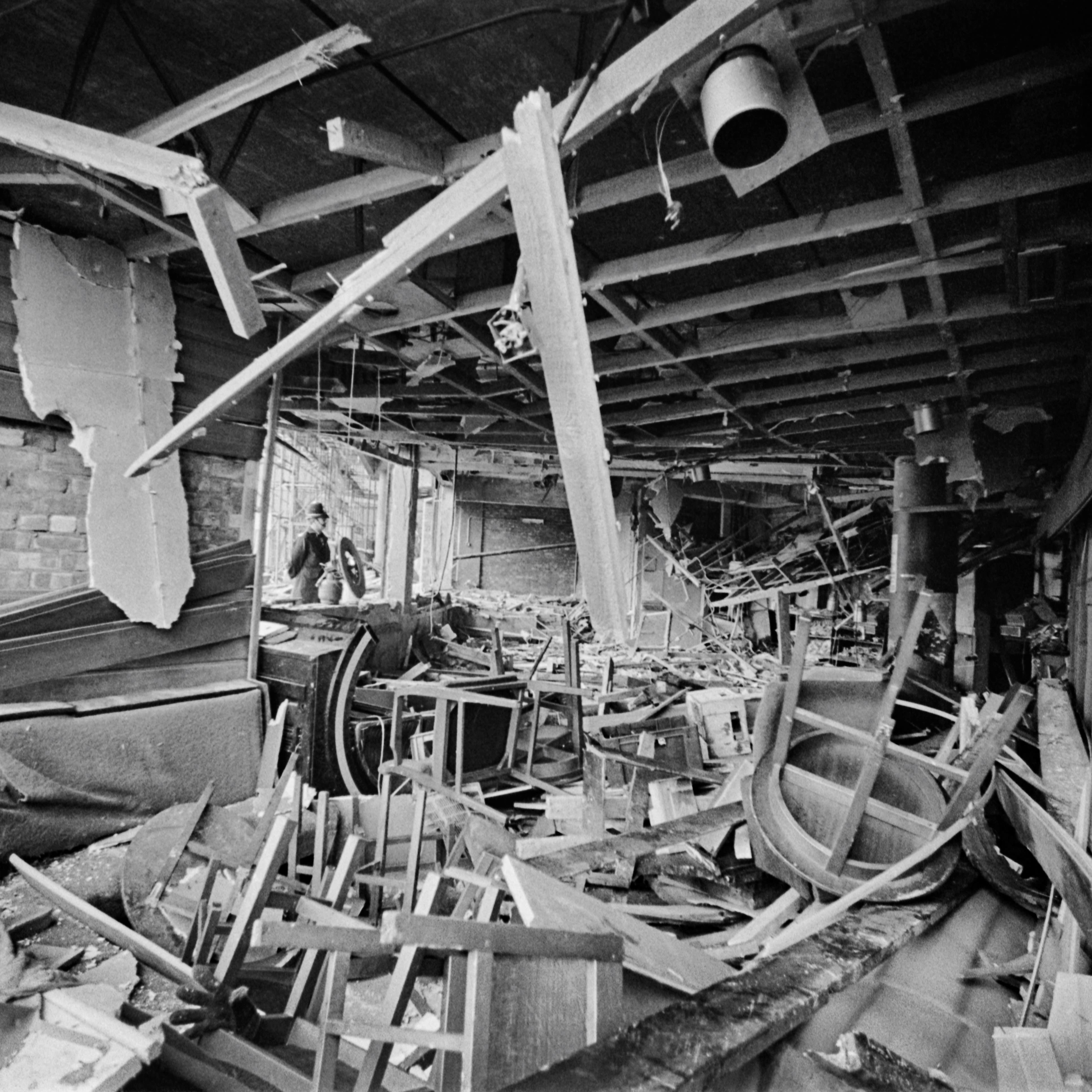 A black and white image of the interior of a pub. There are chairs and beams covering the floor, with wood and pieces of wood hanging down.