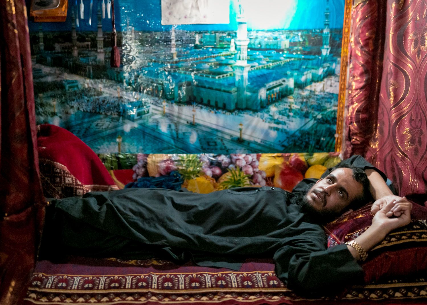 An inmate lies back in his bunk bed decorated by red drapes and a large poster of Medina