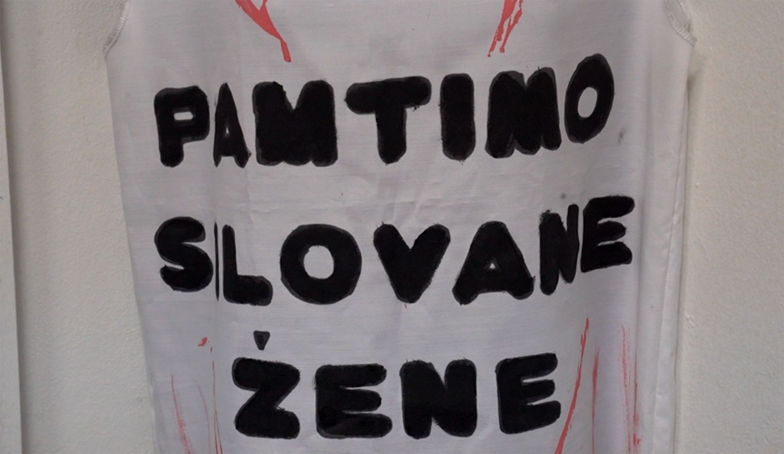 Beli platneni transparent na kome debelim crnim slovima latinicom piše "PAMTIMO SILOVANE ŽENE", sa nekoliko crvenih tragova po platnu