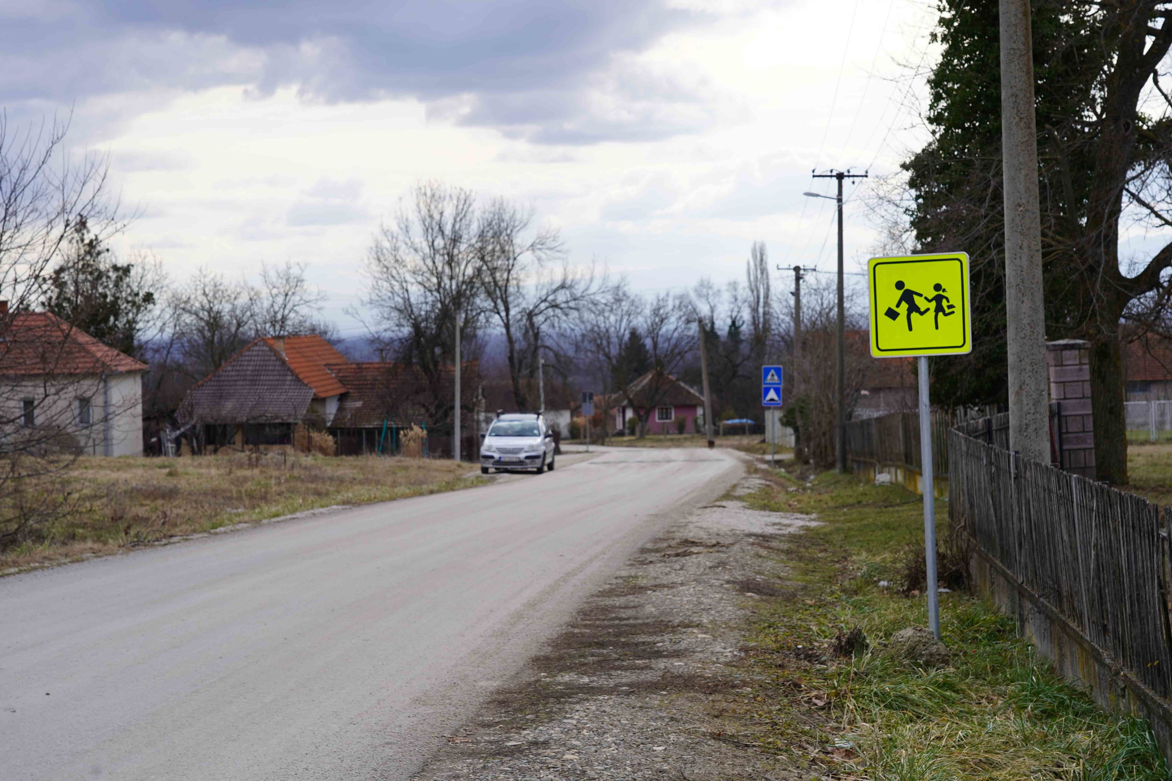 Selo, put, sa desne strane je upadljivi saobraćajni znak za školu koji označava da deca možda pretrčavaju put ovde.