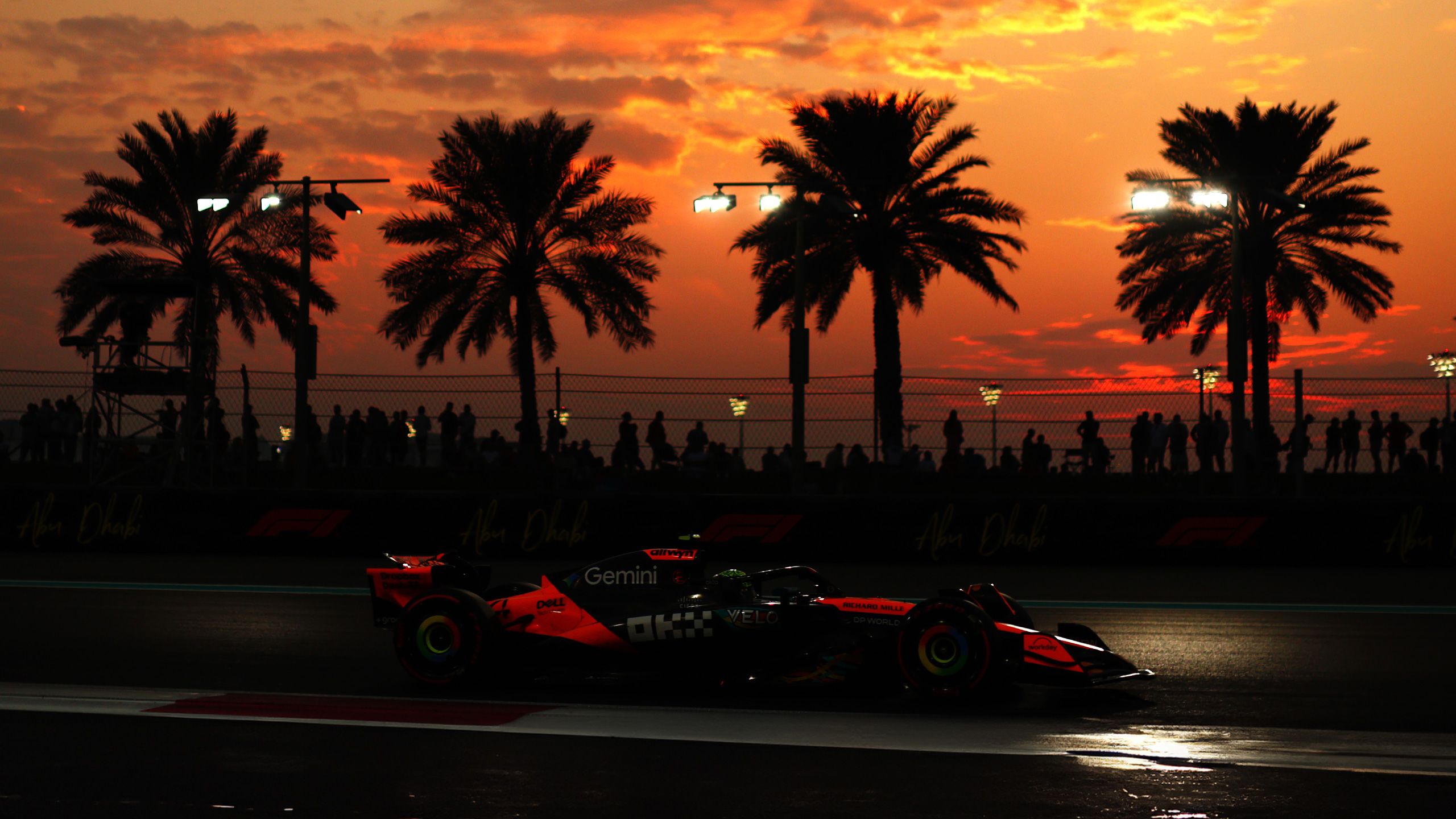 Lewis hamilton Formula 1 driver in a red racing suit standing on stage during an F1 event, with a race car and two other individuals in the background under bright spotlights