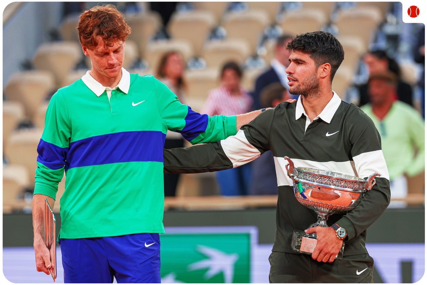 Carlos Alcaraz holds the French Open trophy while consoling Jannik Sinner on a clay court during the award ceremony.