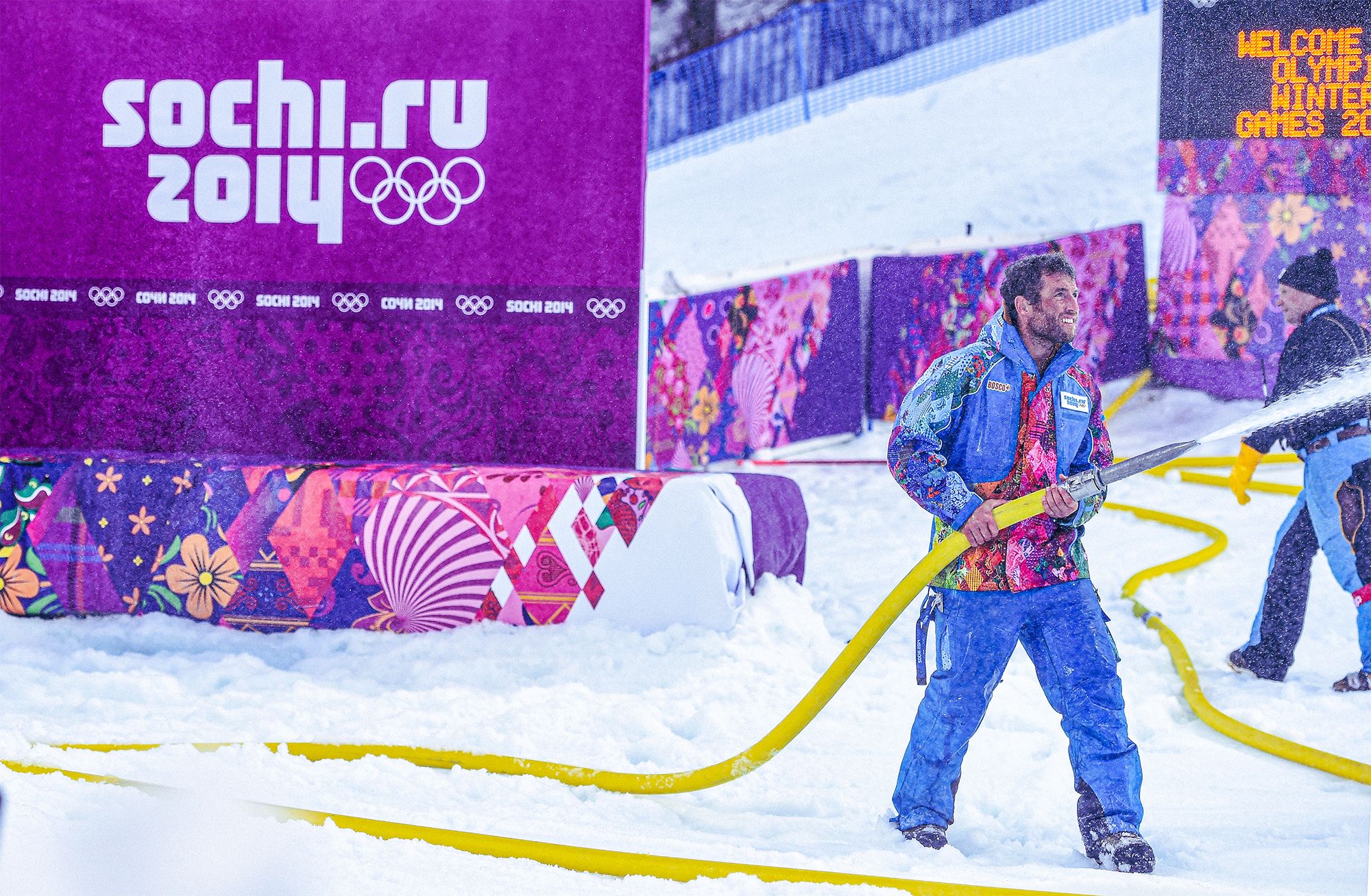 Worker in brightly colored Sochi 2014 Winter Olympics uniform using a hose to spray snow onto a competition area, with purple event branding panels and snowy ground surrounding the scene