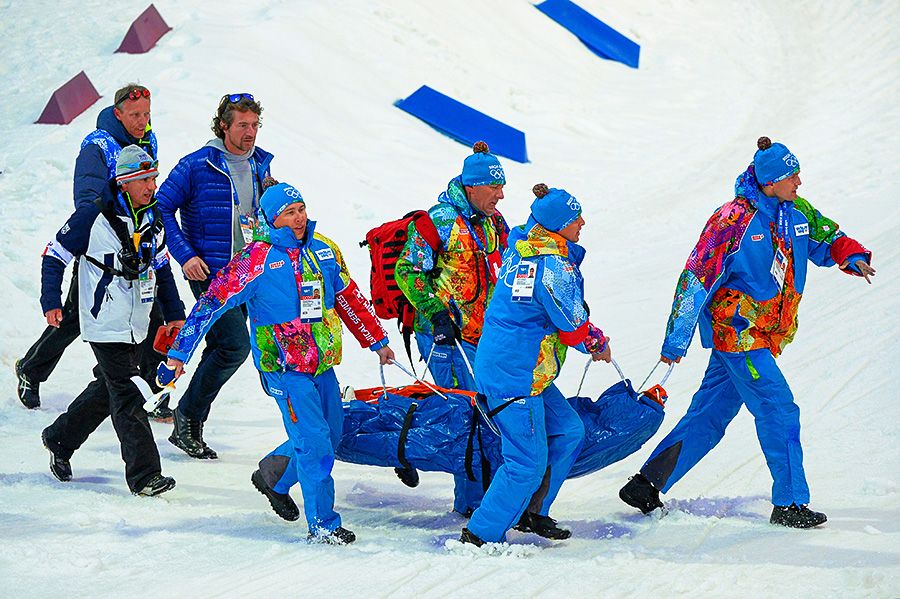 Rescue team in colorful Sochi 2014 Winter Olympics uniforms carrying an injured athlete on a stretcher across a snowy slope, accompanied by support staff walking alongside them