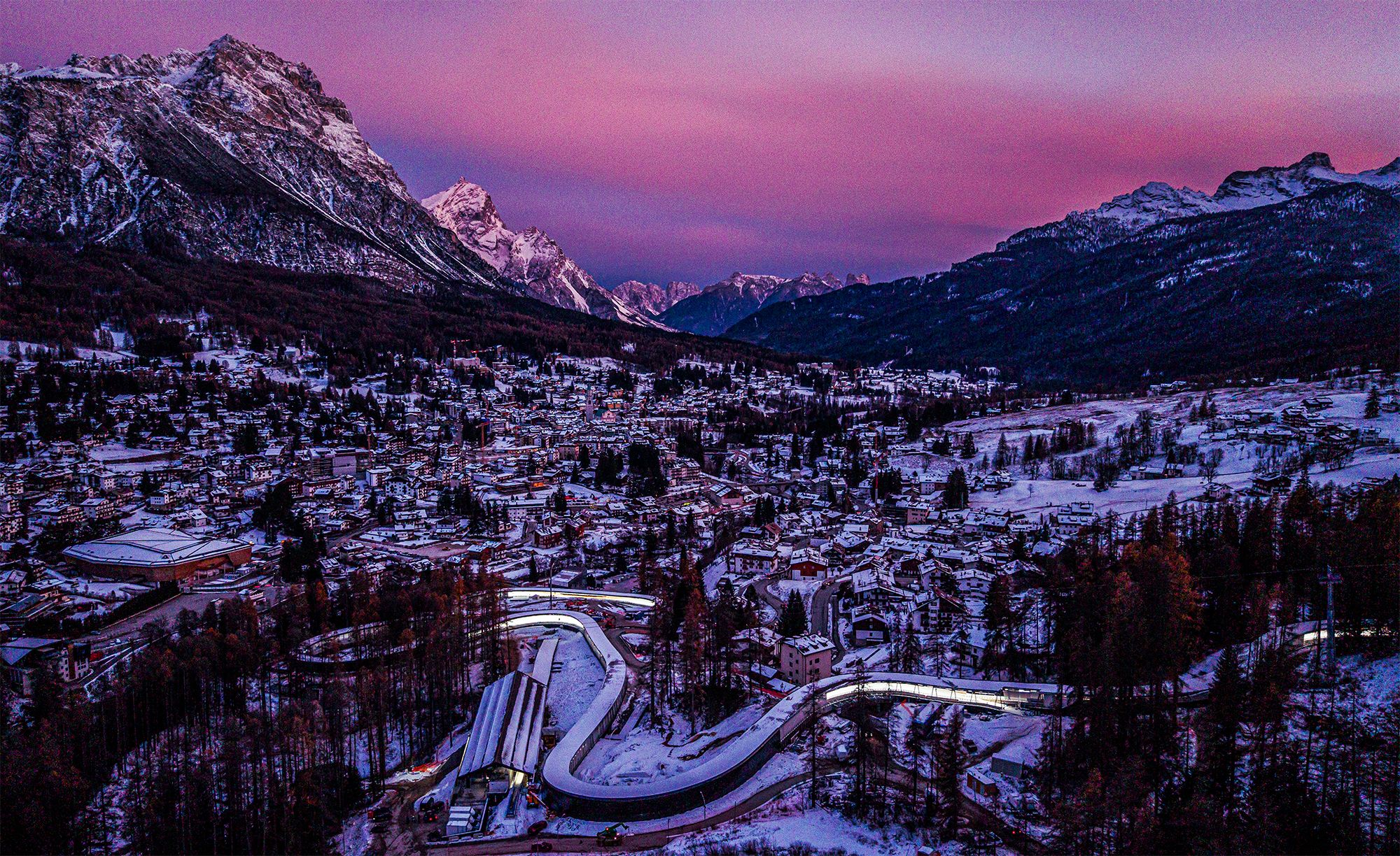 Aerial view of Cortina d’Ampezzo at dusk, with snow‑covered buildings in the valley, a winding illuminated bobsled track in the foreground, and dramatic mountain peaks glowing pink and purple under a sunset sky
