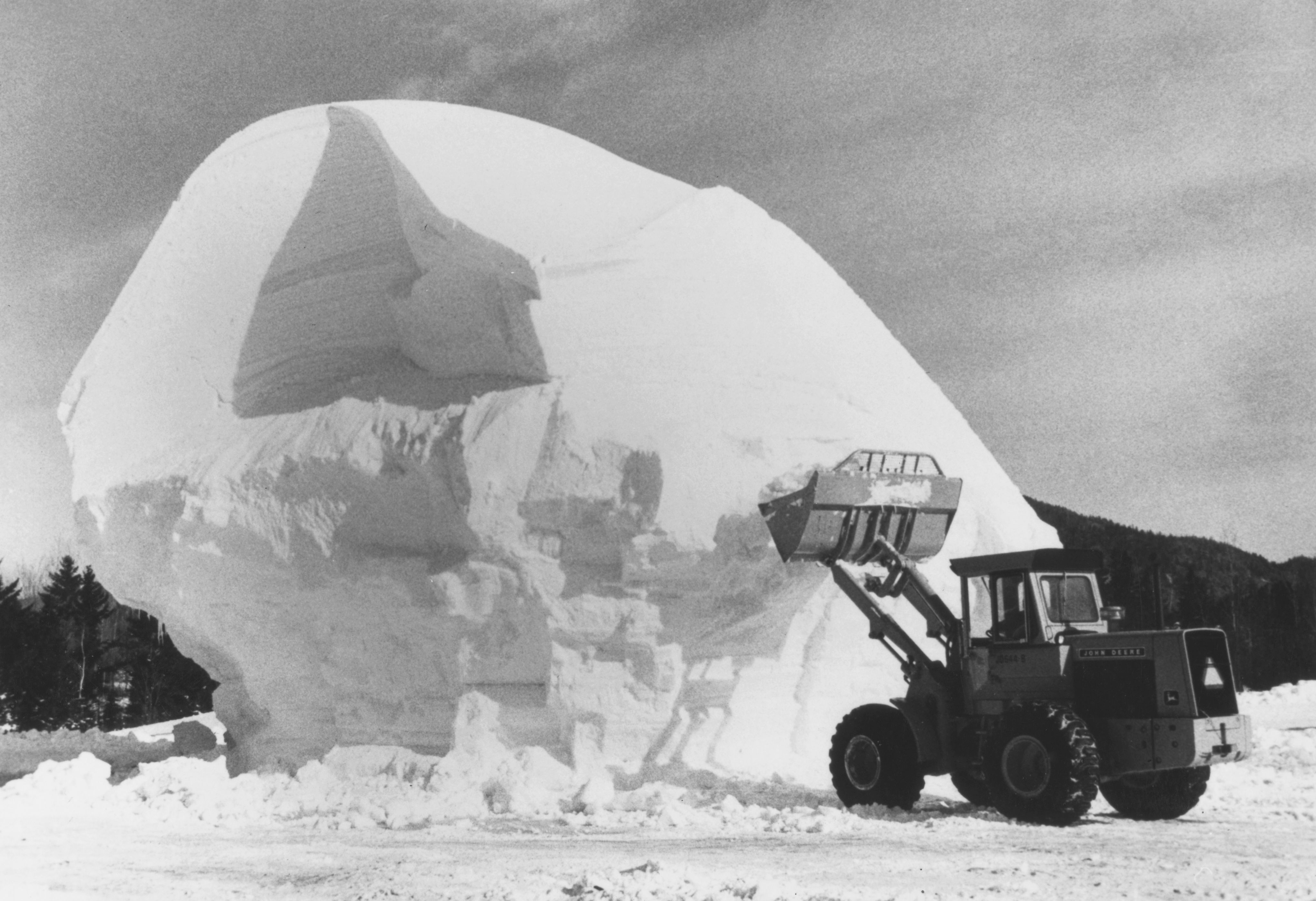 Black‑and‑white photo of a large front‑loader tractor shaping a massive mound of compacted snow, with trees and mountains in the background
