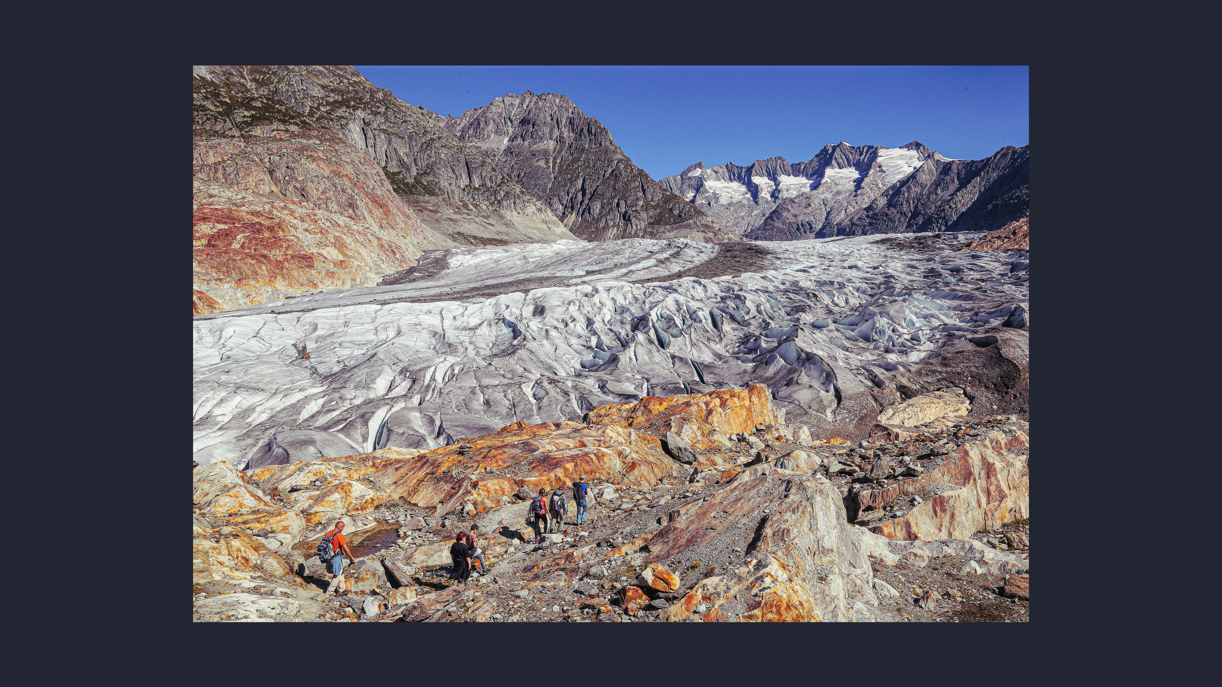 Group of hikers walking across rocky terrain beside a glacier, surrounded by steep mountains and large patches of exposed ice under a clear blue sky