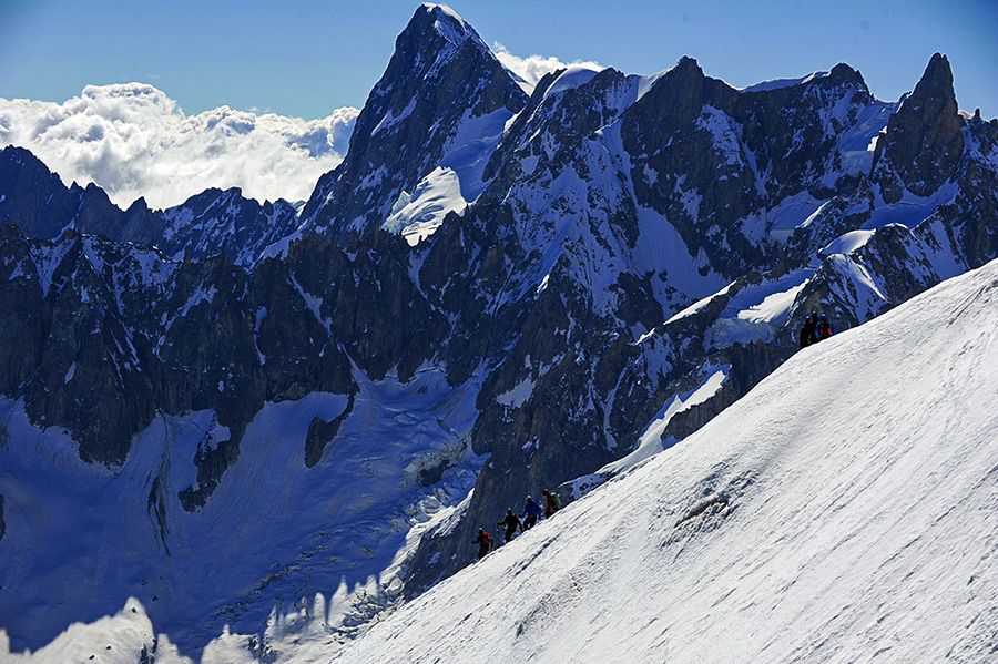 Climbers ascending a steep, snow‑covered slope in the Alps, with dramatic jagged mountain peaks rising behind them and clouds filling the distant valley