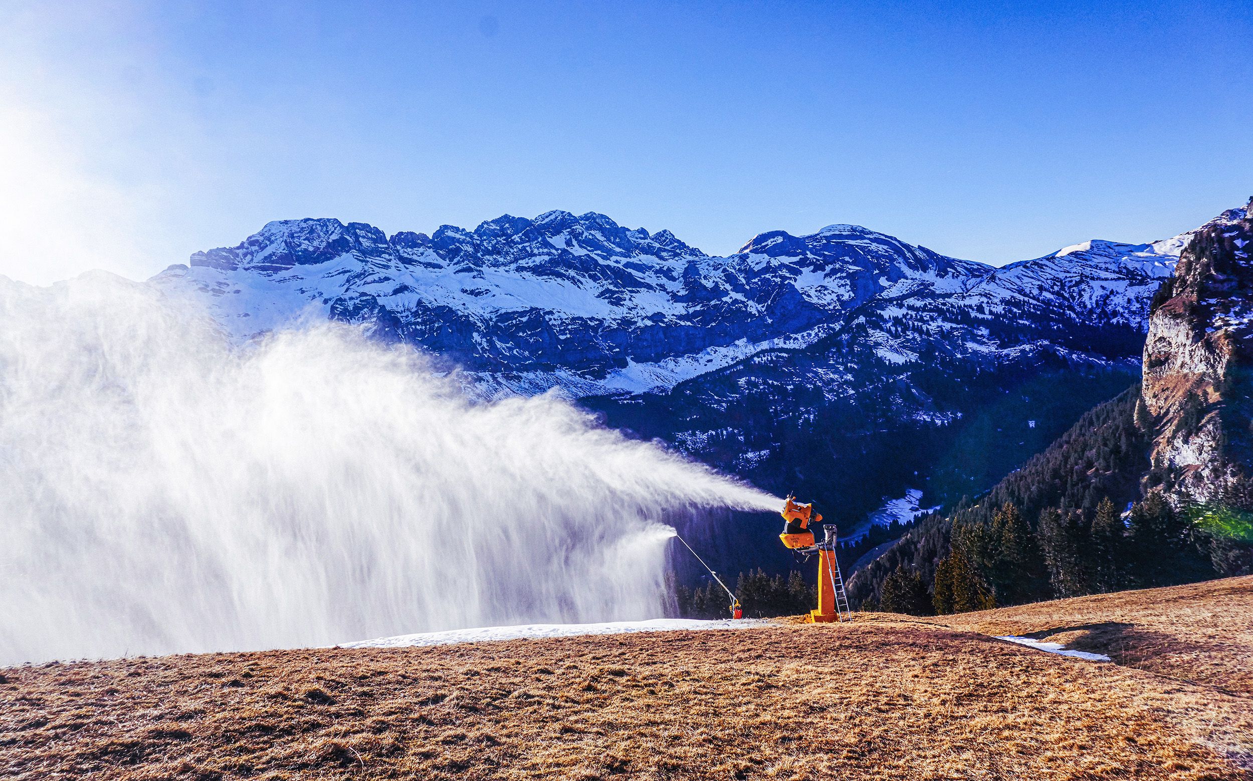 A snowmaking machine sprays a large plume of artificial snow across a mostly brown ski slope, with snow‑covered mountains and a clear blue sky in the background