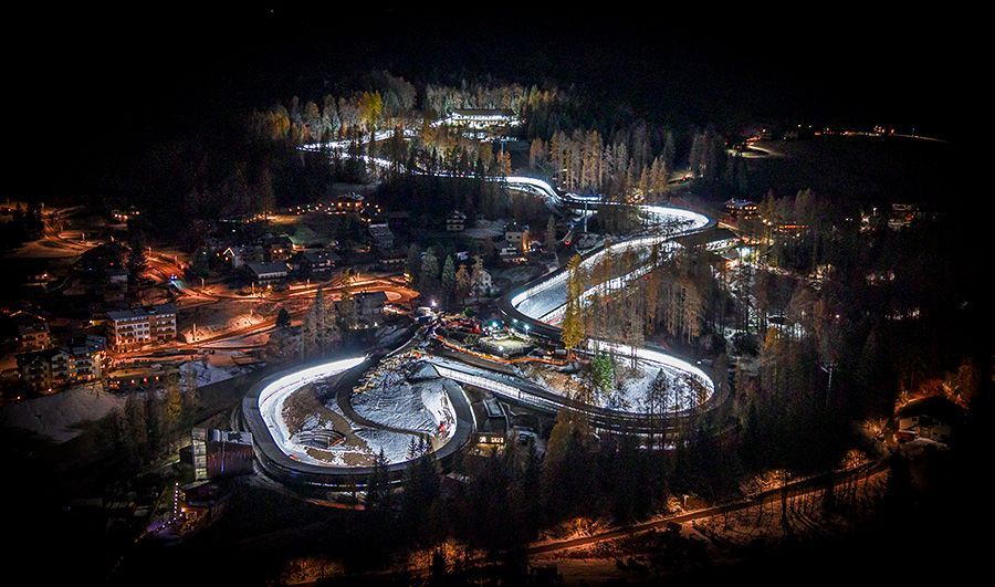 Aerial night view of a brightly lit bobsled and luge track winding through a snowy forested landscape, with nearby buildings and trees illuminated against the darkness