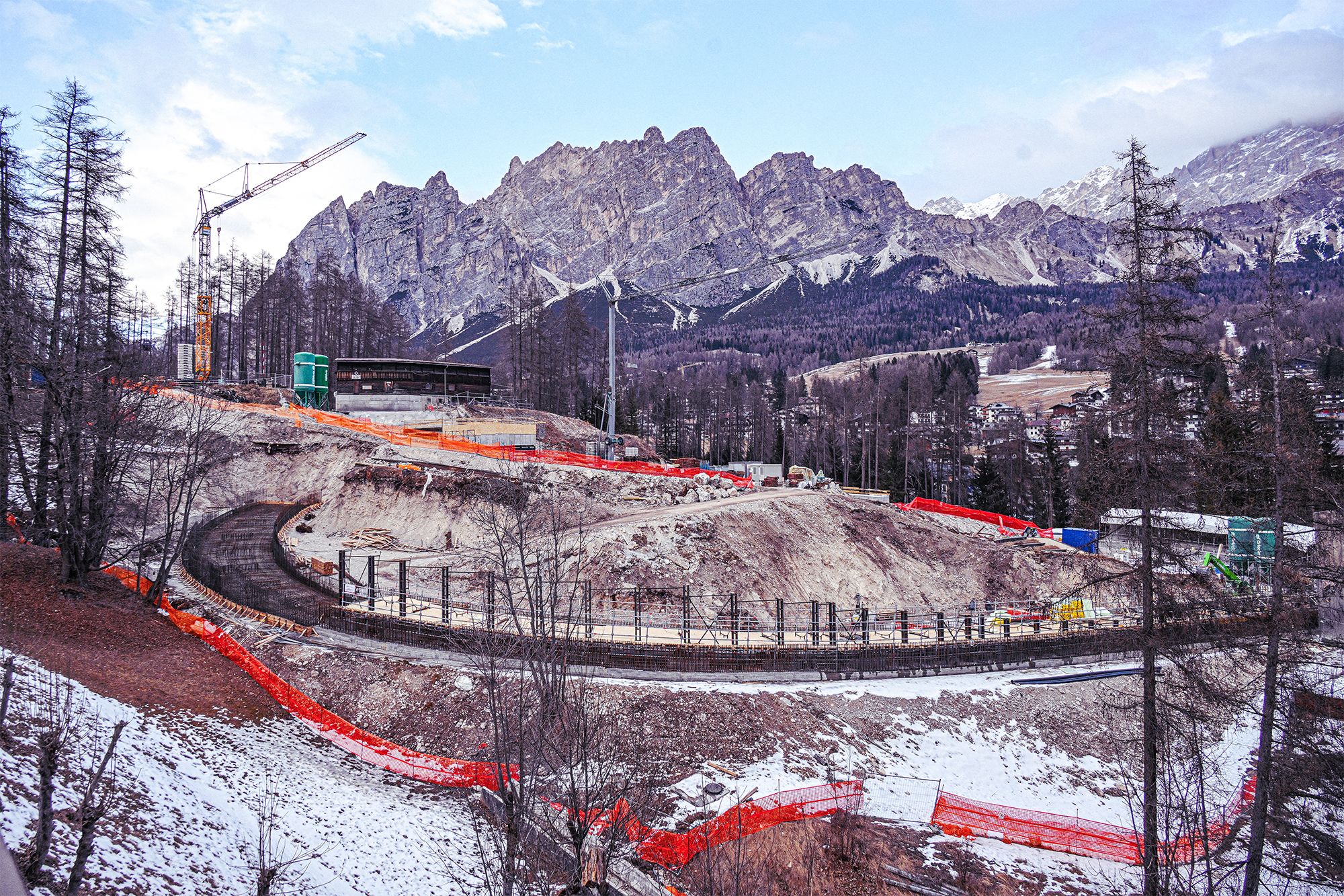 Construction site of a bobsled or luge track in a mountain valley, with winding concrete sections, cranes, orange safety fencing, and snow‑dusted ground beneath rugged peaks