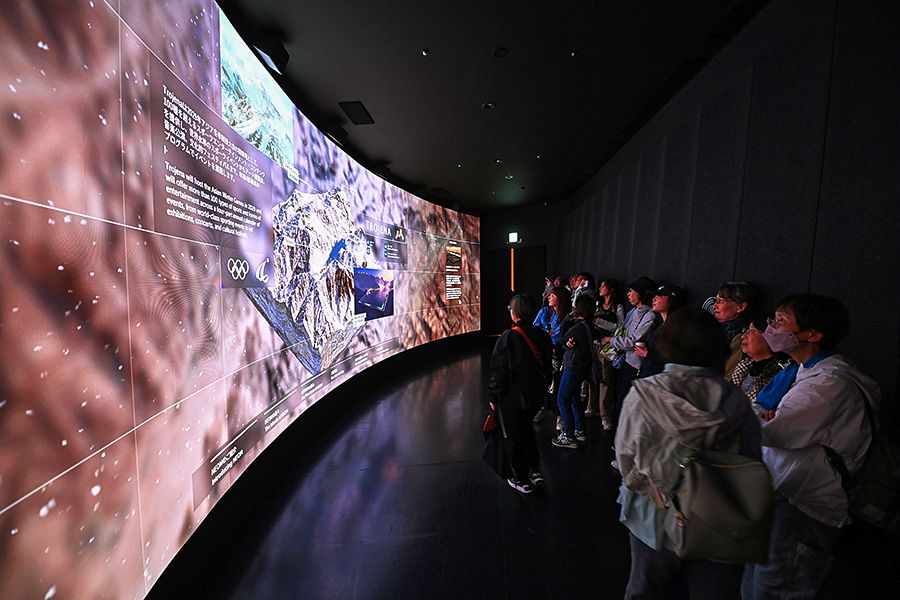 Visitors standing in a dark exhibition space viewing a large curved digital screen displaying proposed Trojema visuals and text