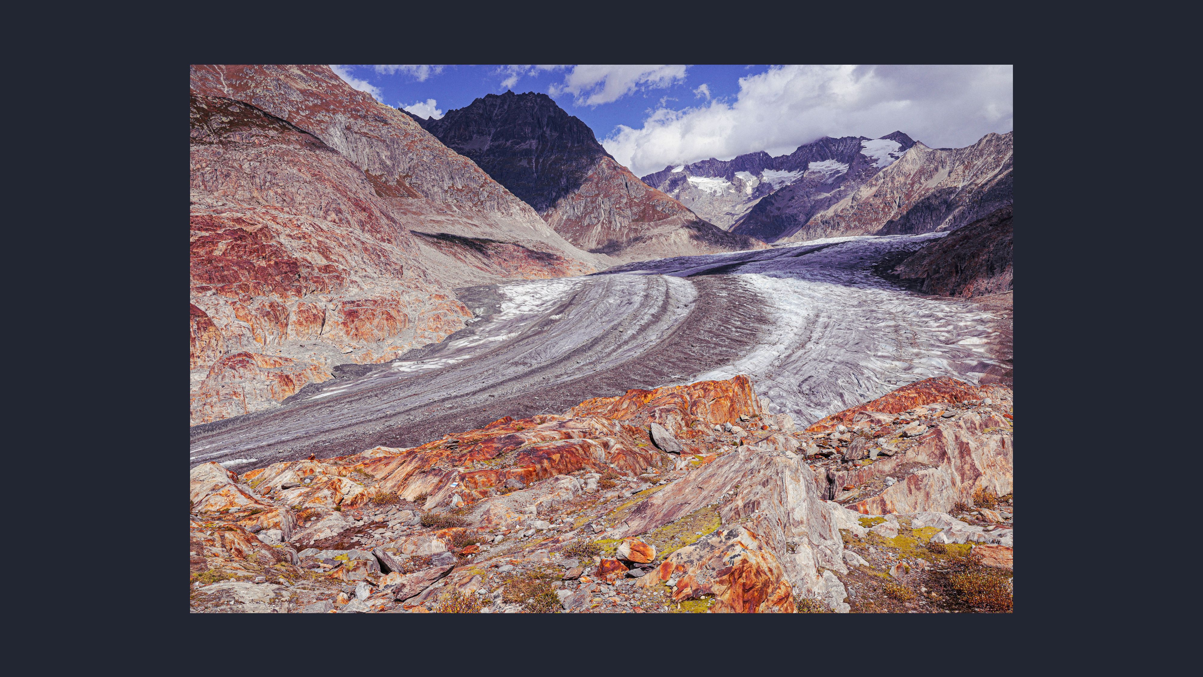 The same view but the glacier is significantly smaller through a rocky mountain valley, with exposed red‑brown rock in the foreground and snow‑covered peaks under a partly cloudy sky