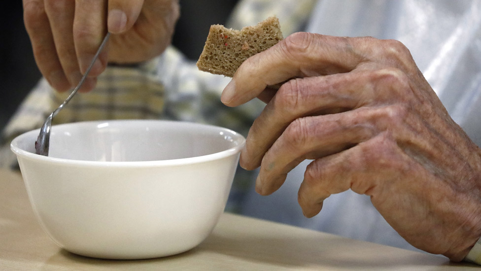 Old person's hand holding a piece of bread and the other hand is holding a spoon over a bowl